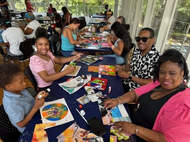 Children and adults sitting at long tables in a bright room, engaging in arts and crafts activities with colorful paper, glue, scissors, and magazines.