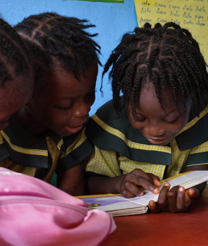 Two young girls with braided hair read a book together in a classroom, wearing green and yellow uniforms.