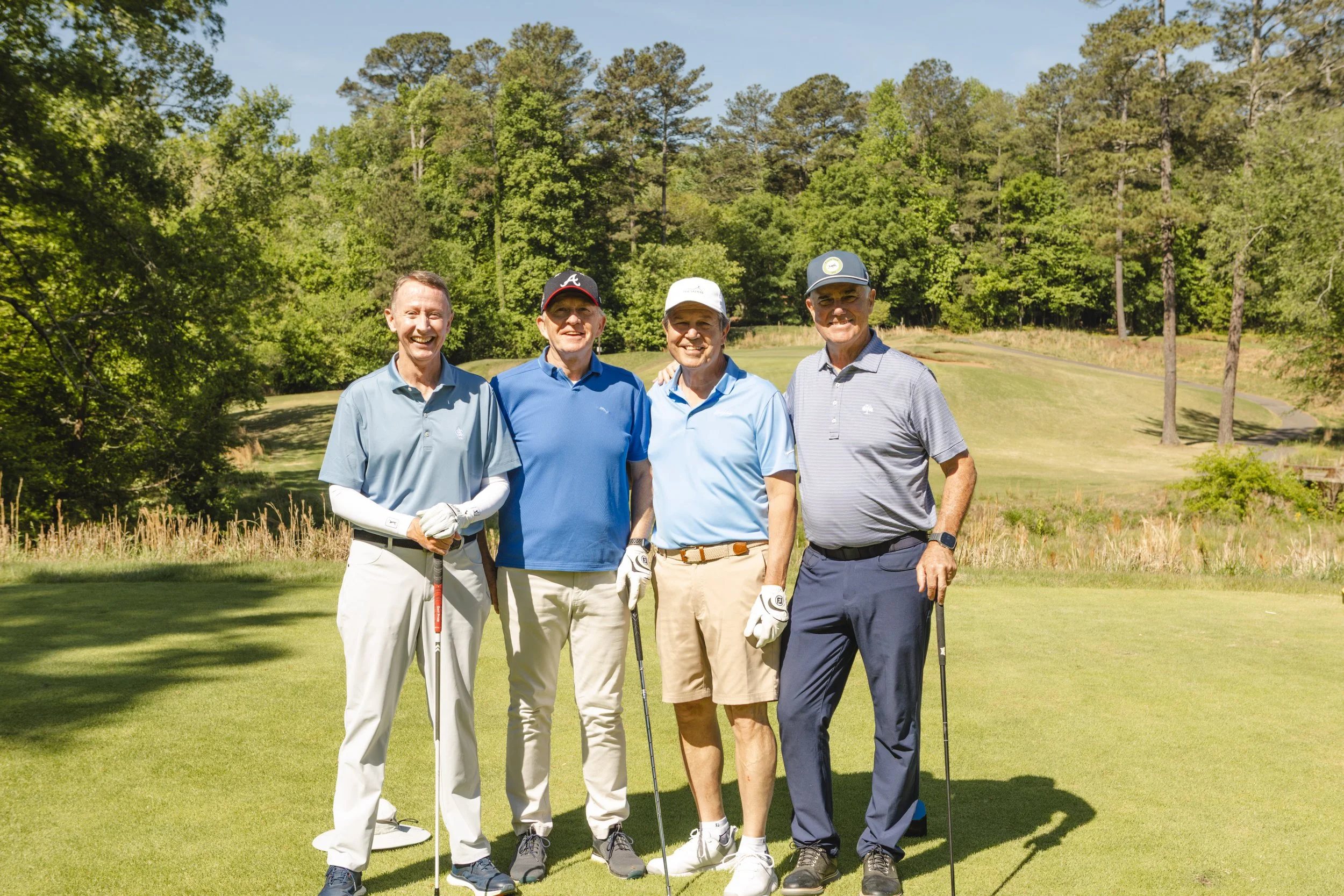Four men standing on a golf course, smiling and posing for a photo, holding golf clubs, with trees and a clear sky in the background.