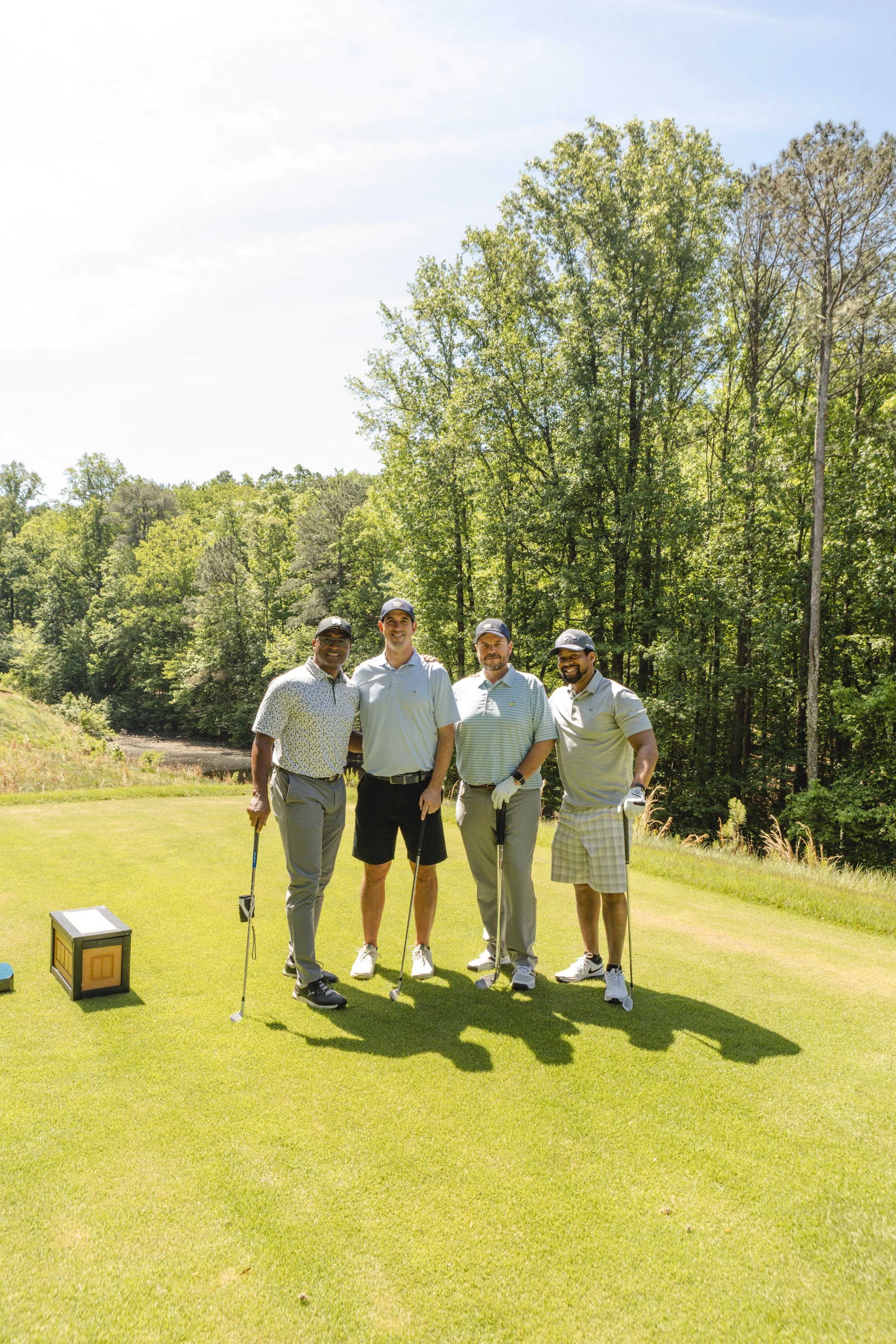 Four men are standing on a golf course, smiling for the camera, with trees in the background and golf clubs in their hands.