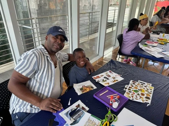 A man and a boy sitting at a craft table with paper cutouts of various animals and insects, smiling at the camera. Other children at similar tables are engaged in craft activities.