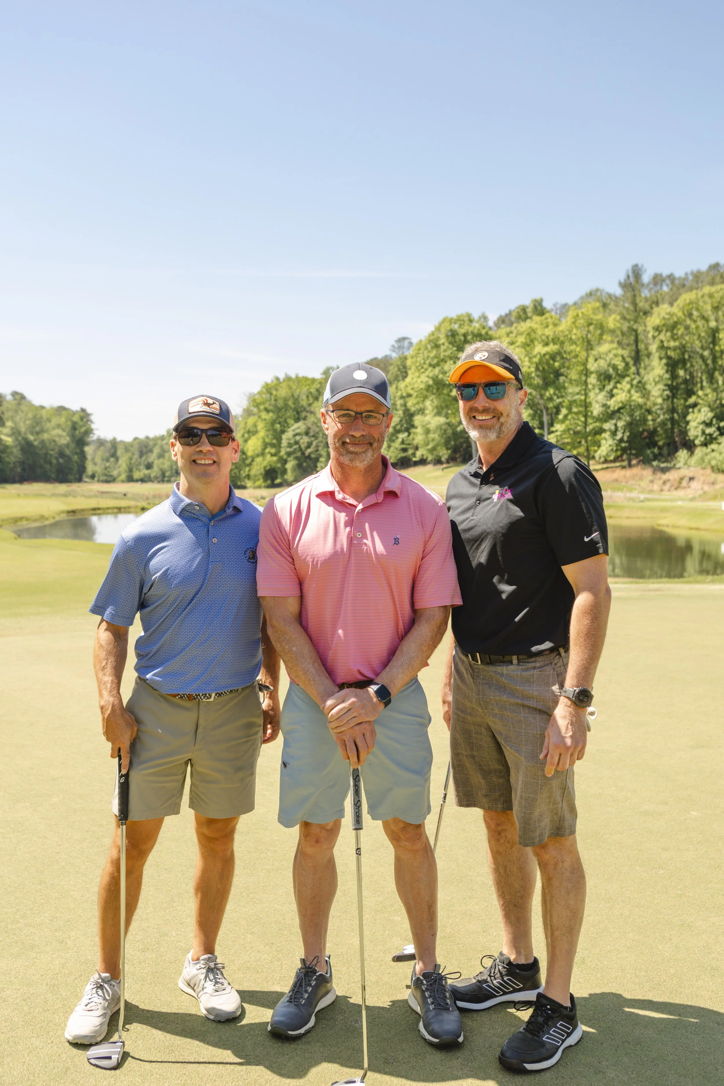 Three middle-aged men standing on a golf course, smiling for the camera, holding golf clubs. They are dressed in golf attire with caps and sunglasses. The background shows a water hazard and lush green trees under a clear blue sky.