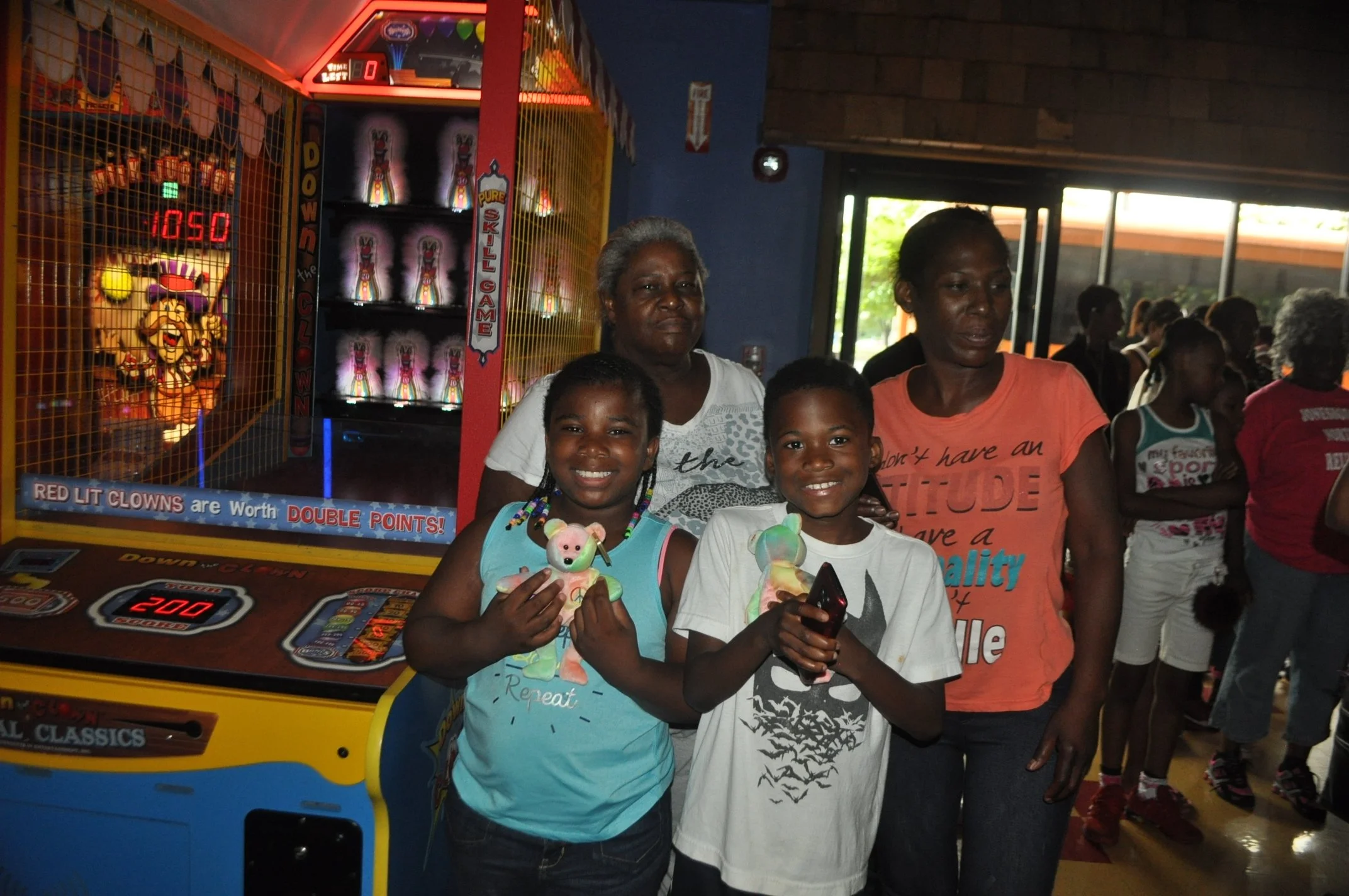 Two smiling girls holding stuffed animals and enjoying arcade games with two women standing behind them at an indoor arcade center.