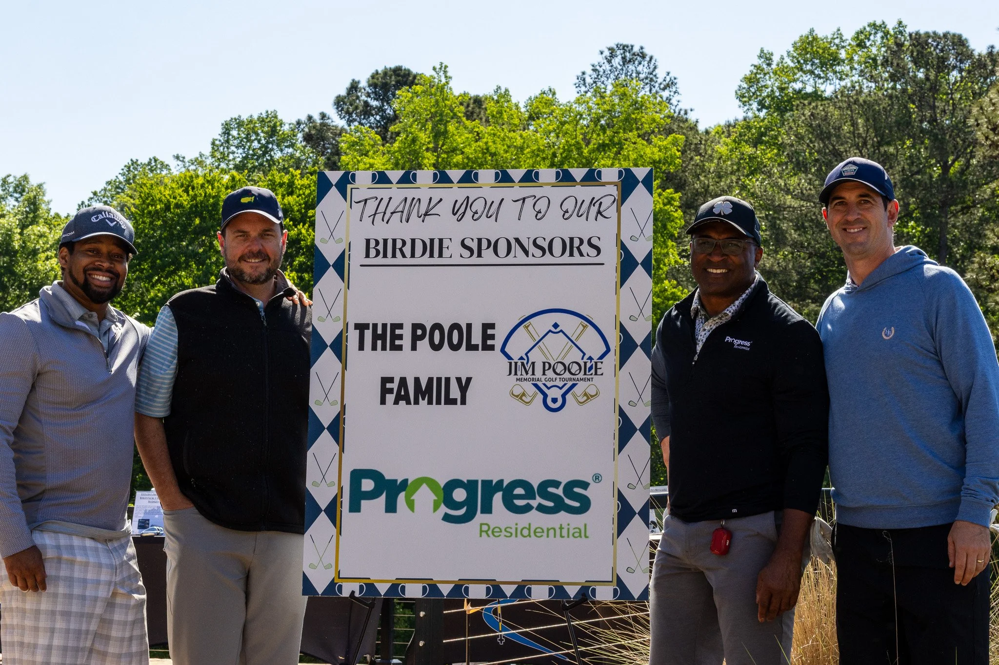 Four men standing outdoors near a sign that thanks sponsors for a golf event, with trees and a clear sky in the background.