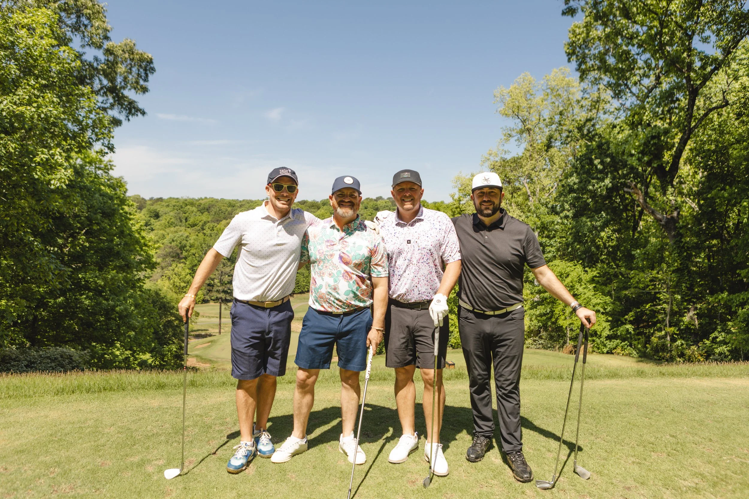 Four men standing together on a golf course, smiling, holding golf clubs, surrounded by lush green trees under a clear blue sky.