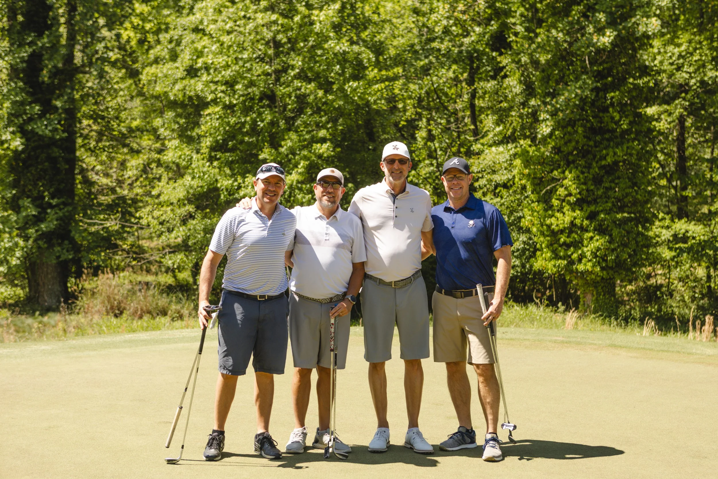 A group of five men standing together on a golf course, holding golf clubs, with trees in the background.