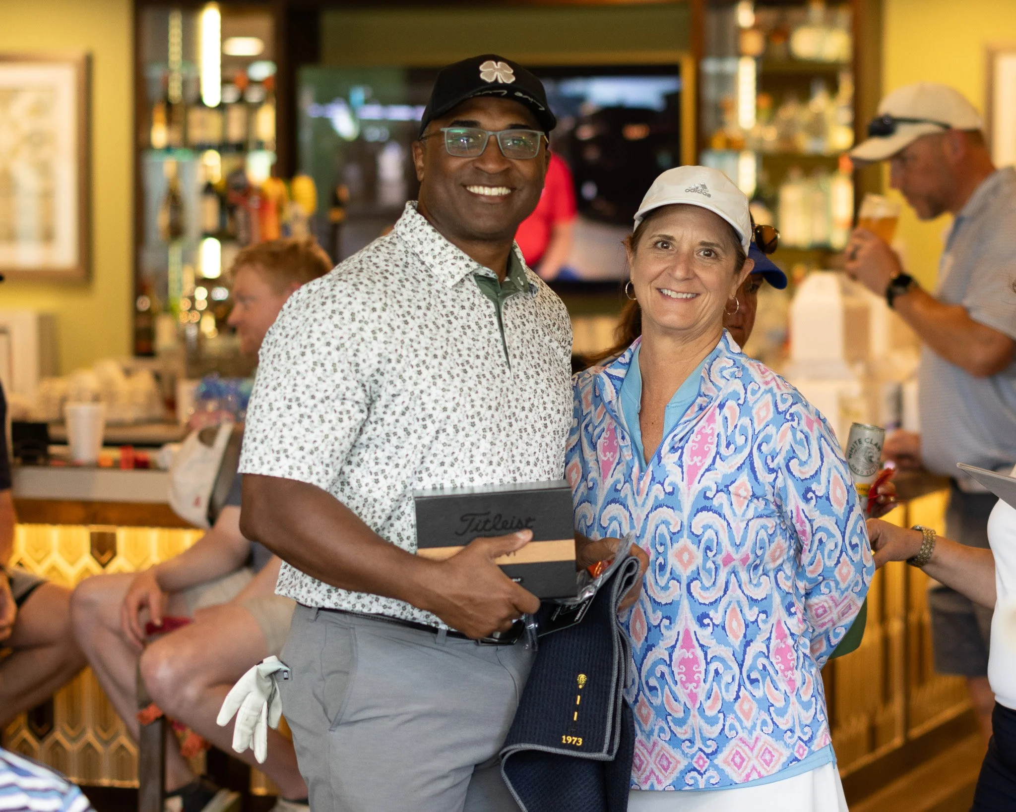 A man and woman smiling at a bar or restaurant. The man is wearing a white floral shirt, gray pants, and glasses and holds a black box. The woman wears a colorful patterned blouse and a white cap. Other people are in the background, some seated and s