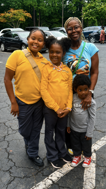 A smiling woman and three children standing together outdoors in a parking lot with cars and trees in the background. The woman has braids and glasses, and the children are wearing casual clothes, two in yellow and one in gray.