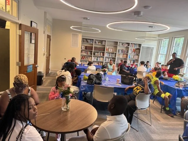 Children sitting at tables during an indoor event in a room with a bookshelf, some adults supervising, and modern circular ceiling lights.