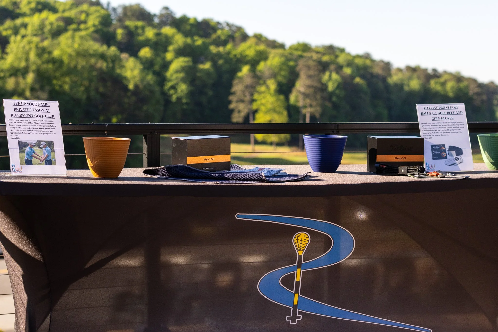 Golf driving range with signs, golf balls, a golf glove, and a golf club on a table, with trees and sunlight in the background.