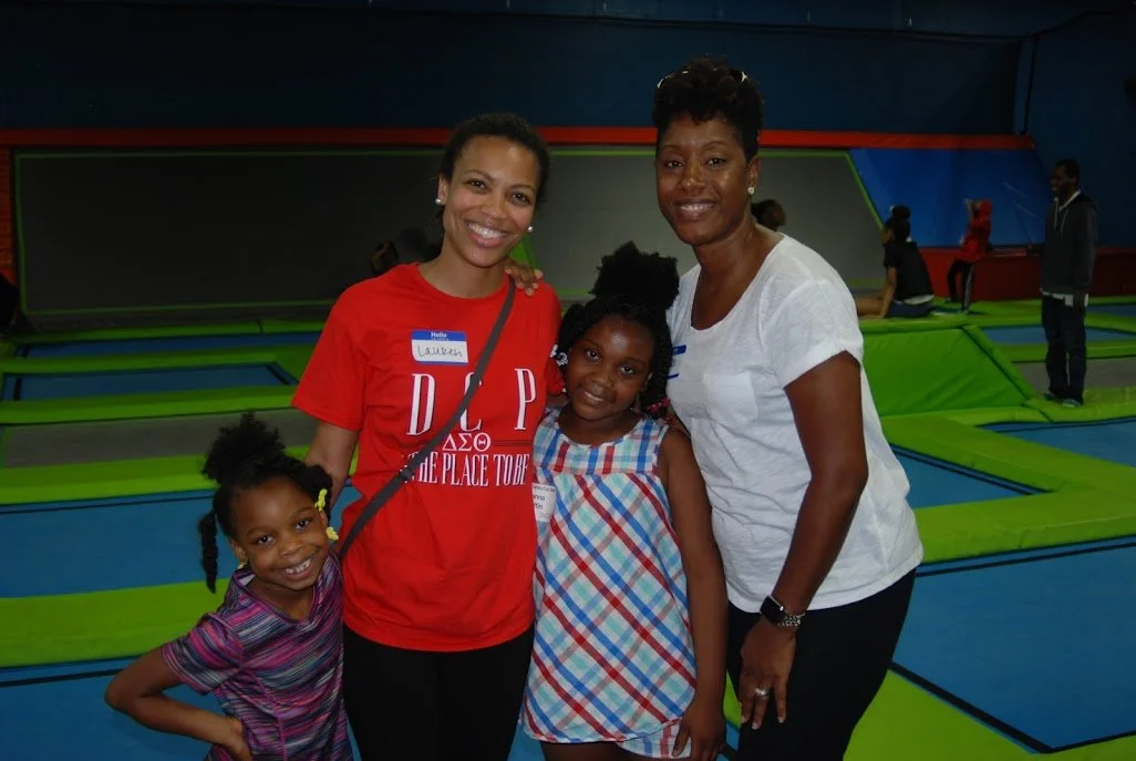 Four females posing inside an indoor trampoline park, with blue and green padded trampolines in the background. Two women and two young girls, all smiling.
