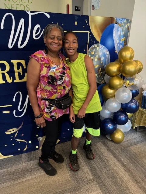 A smiling older woman and a young girl standing together at a celebration, with colorful balloons and a decorative backdrop in the background.