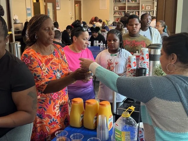 People at a community event with a table of food and drinks, some people are serving and others are waiting.