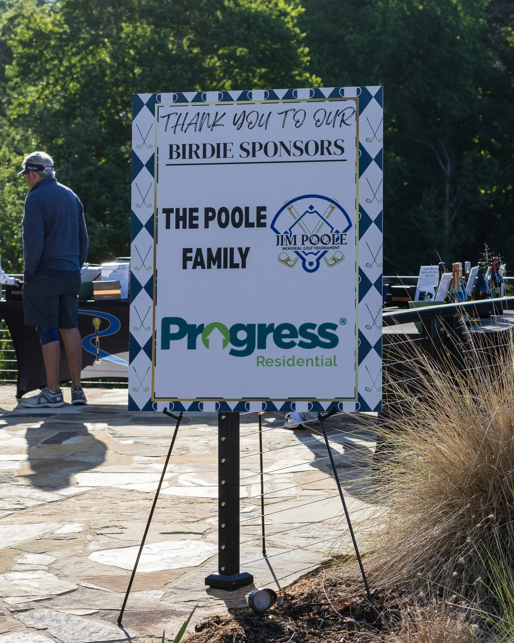 Outdoor event sign thanking sponsors for the Jim Poole Memorial Golf Tournament, with trees in the background and a person in sunglasses and a cap nearby.
