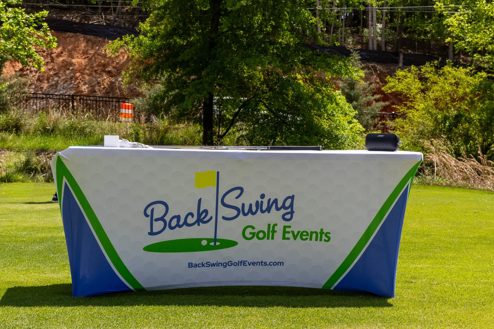 A table covered with a cloth displaying the logo for Back Swing Golf Events at an outdoor golf course, with trees and a rocky hillside in the background.