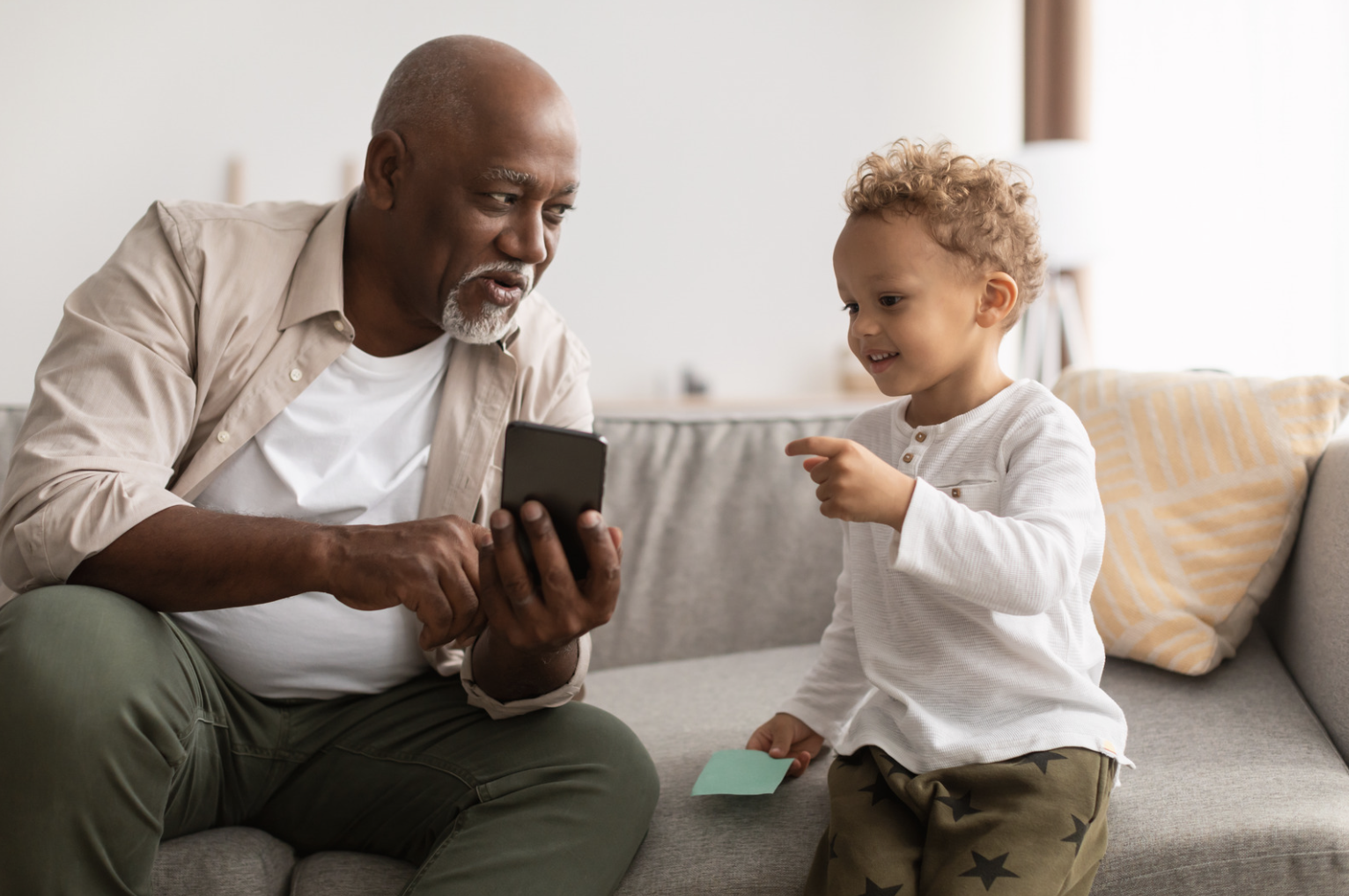 An elderly man and a young boy sitting on a gray sofa in a living room, engaging with a smartphone and a small piece of paper, with a yellow and white pillow in the background.