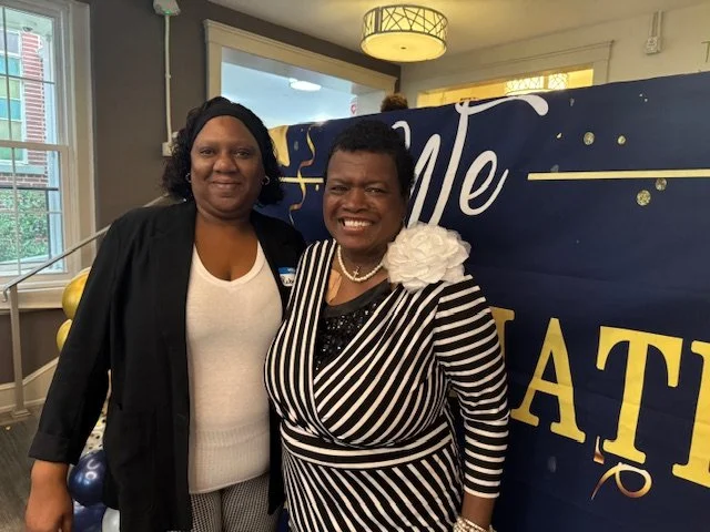 Two women standing together indoors in front of a blue ribbon with yellow lettering, smiling at the camera. One woman wears a black blazer and white shirt, the other wears a black and white striped dress with a large white flower accessory.