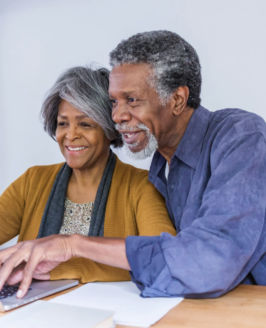 An elderly couple smiling and looking at a laptop together at a table.