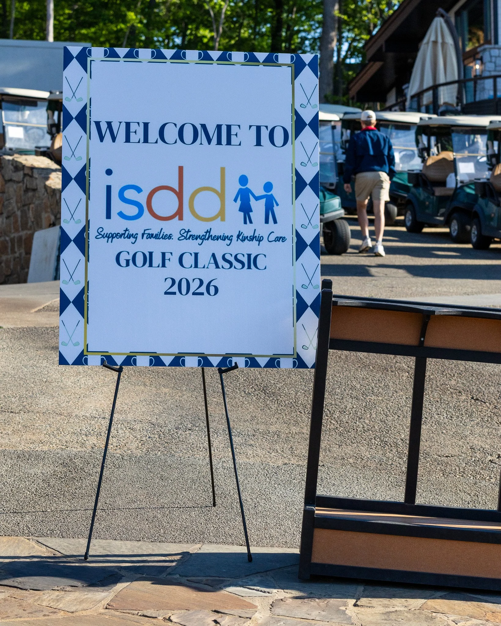 A signboard welcoming guests to the 2026 GOLF CLASSIC event, supporting families and strengthening kinship care, with a decorative border of crossed golf clubs. In the background, a person is walking near golf carts and a building with outdoor seatin