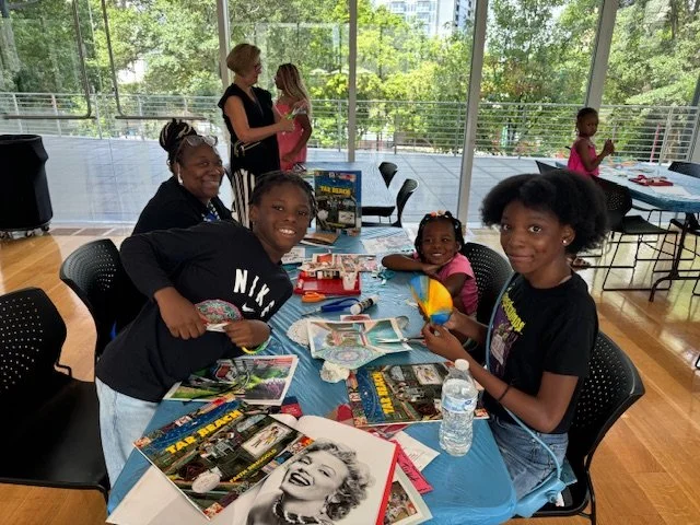 Children and adults gathered around a table with crafts and magazines, in a room with large windows overlooking trees.