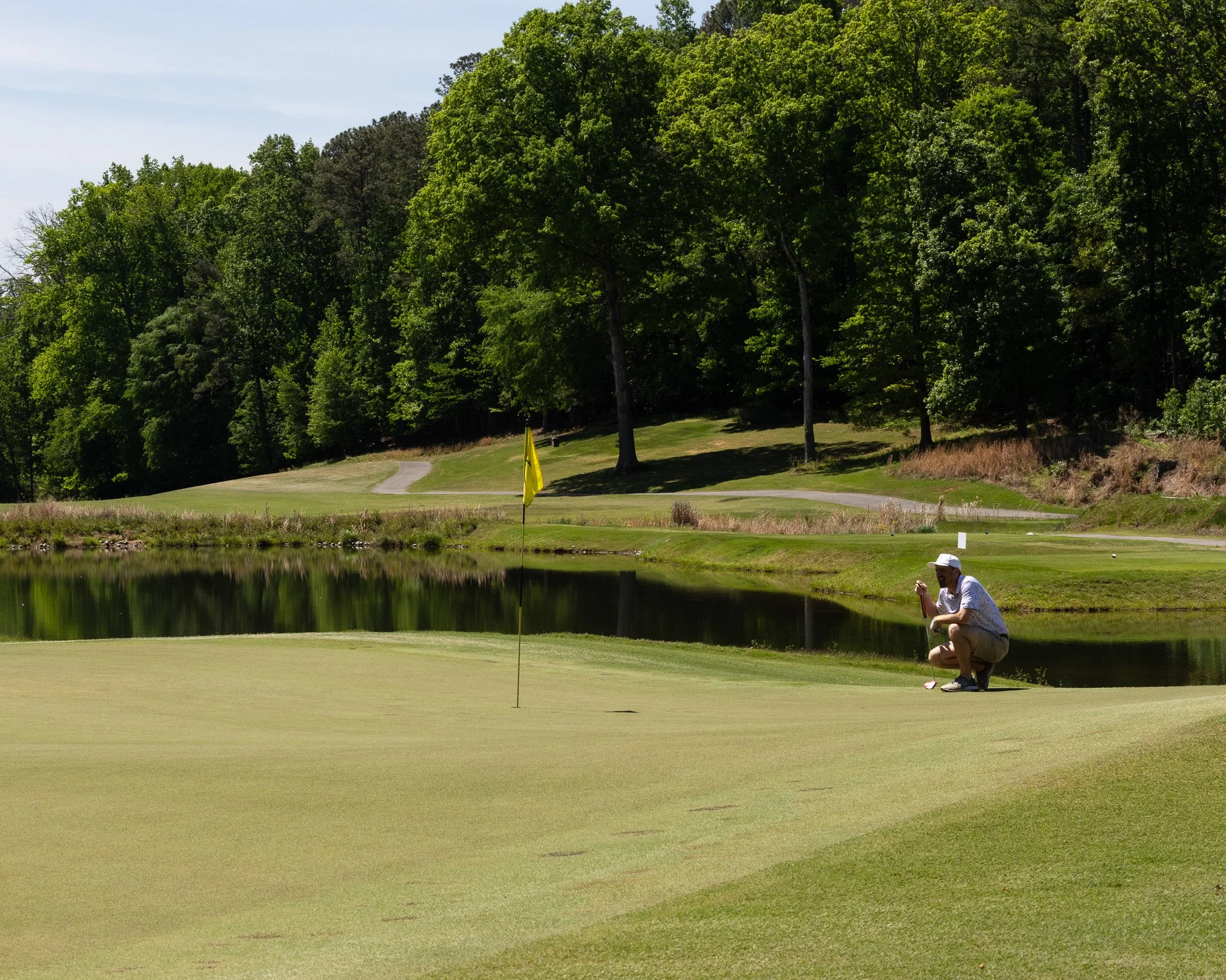 A man kneeling on a golf course green near a water hazard, with trees in the background, preparing to putt towards the hole with a yellow flag.