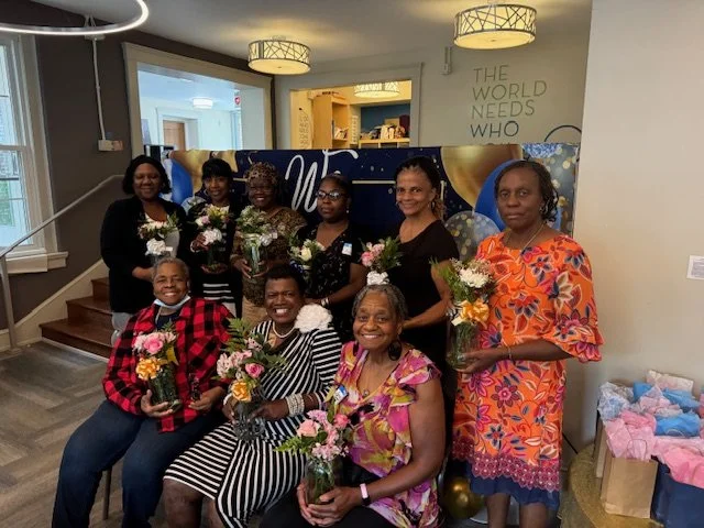 Nine women gathered together, each holding flowers, in a room with a banner and celebratory decorations.