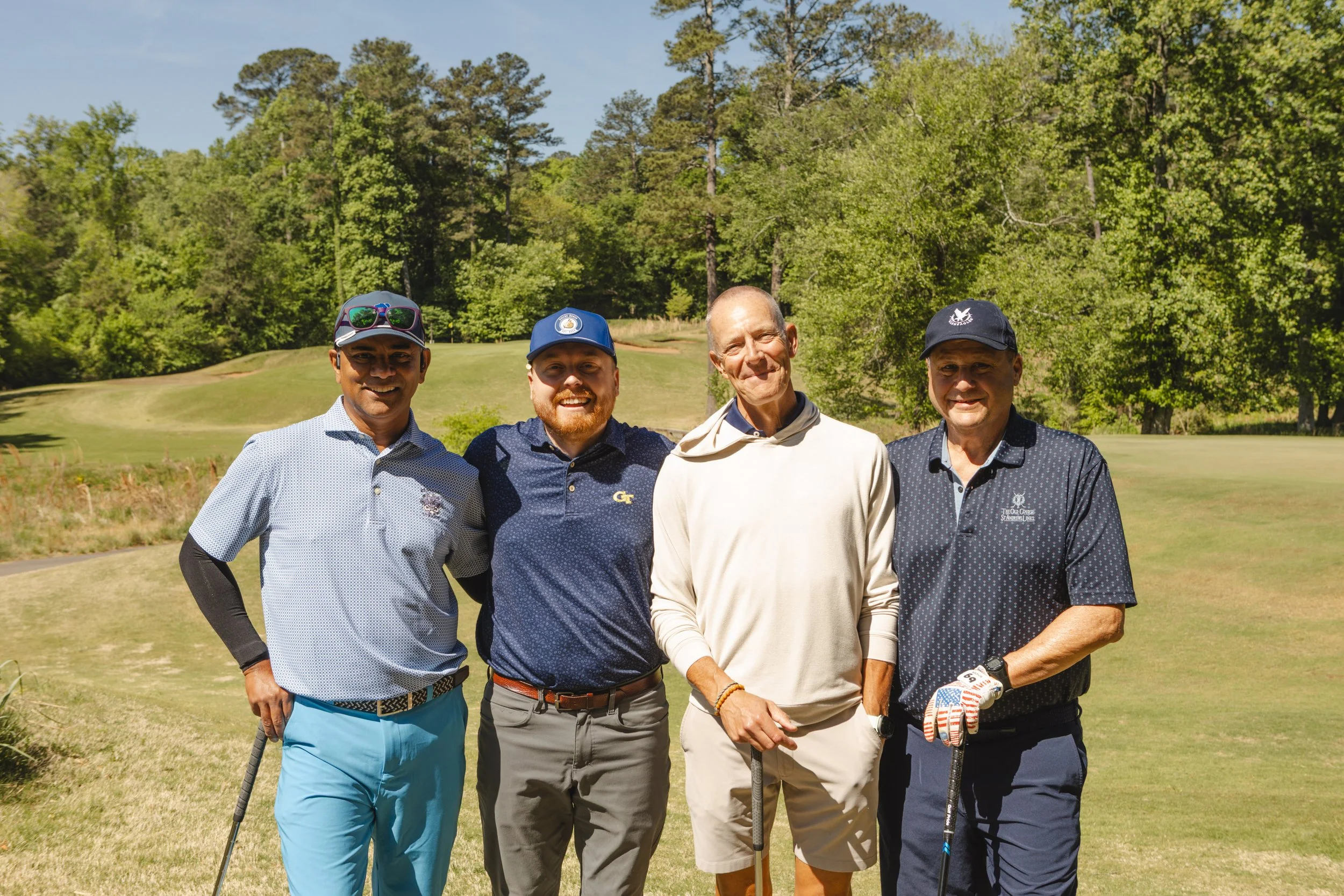 Four men standing together on a golf course, smiling at the camera, surrounded by green trees and grass, with clear blue sky overhead.