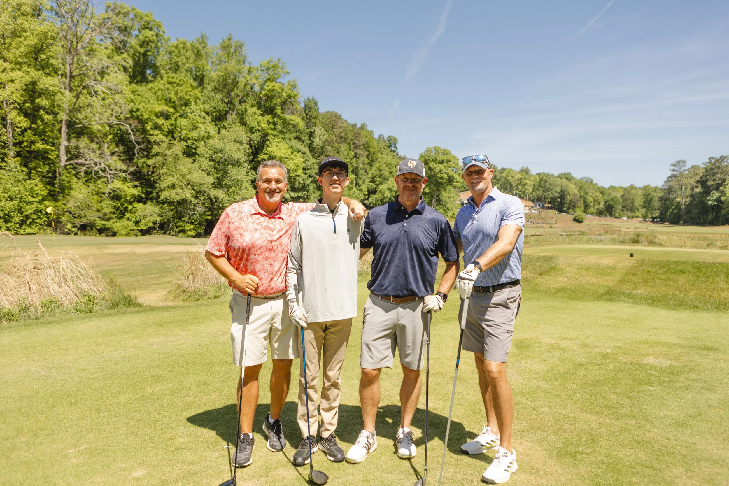 Four men standing on a golf course, smiling, with golf clubs. They are dressed in golf attire, and there are trees and a blue sky in the background.