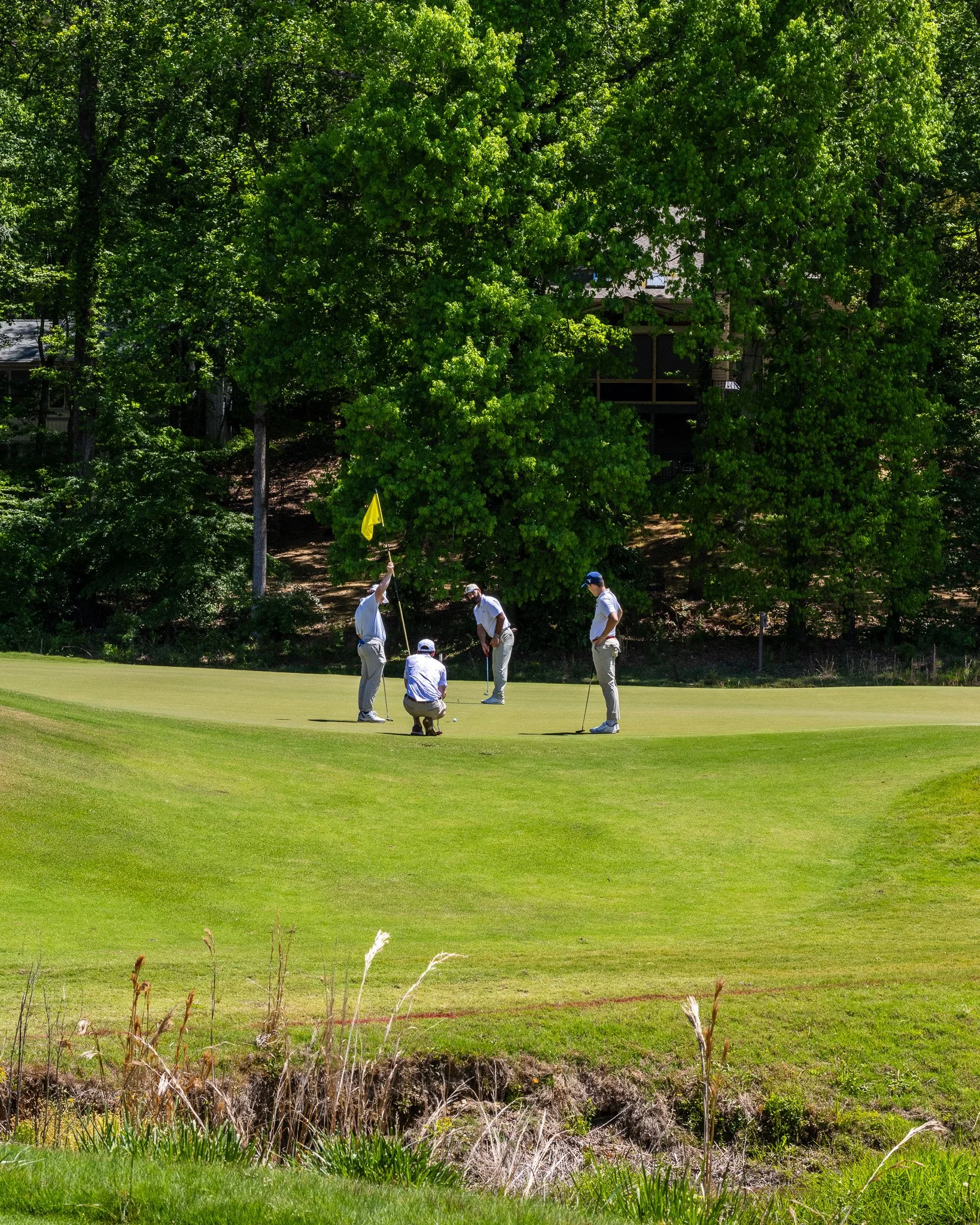 Golf course with three men playing near a green area, with dense trees and a house with a porch in the background.