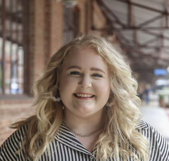 A young woman with blonde, curly hair smiling outdoors near a brick building.