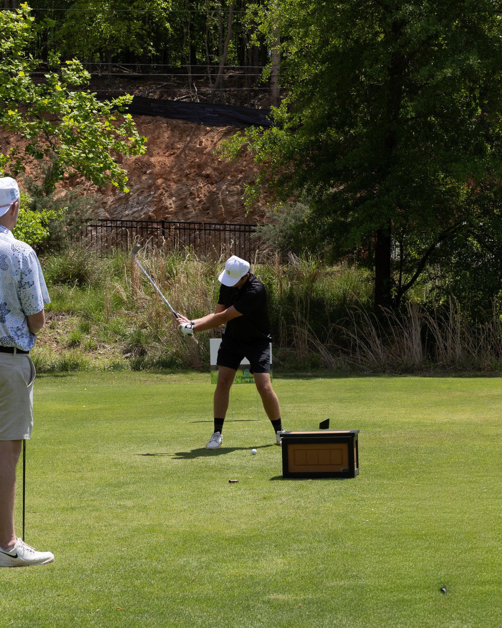 A person dressed in dark clothing and a white cap is preparing to hit a golf ball on a green golf course, with a black box and tee nearby; two other people are partially visible to the left, and trees and a dirt slope form the background.