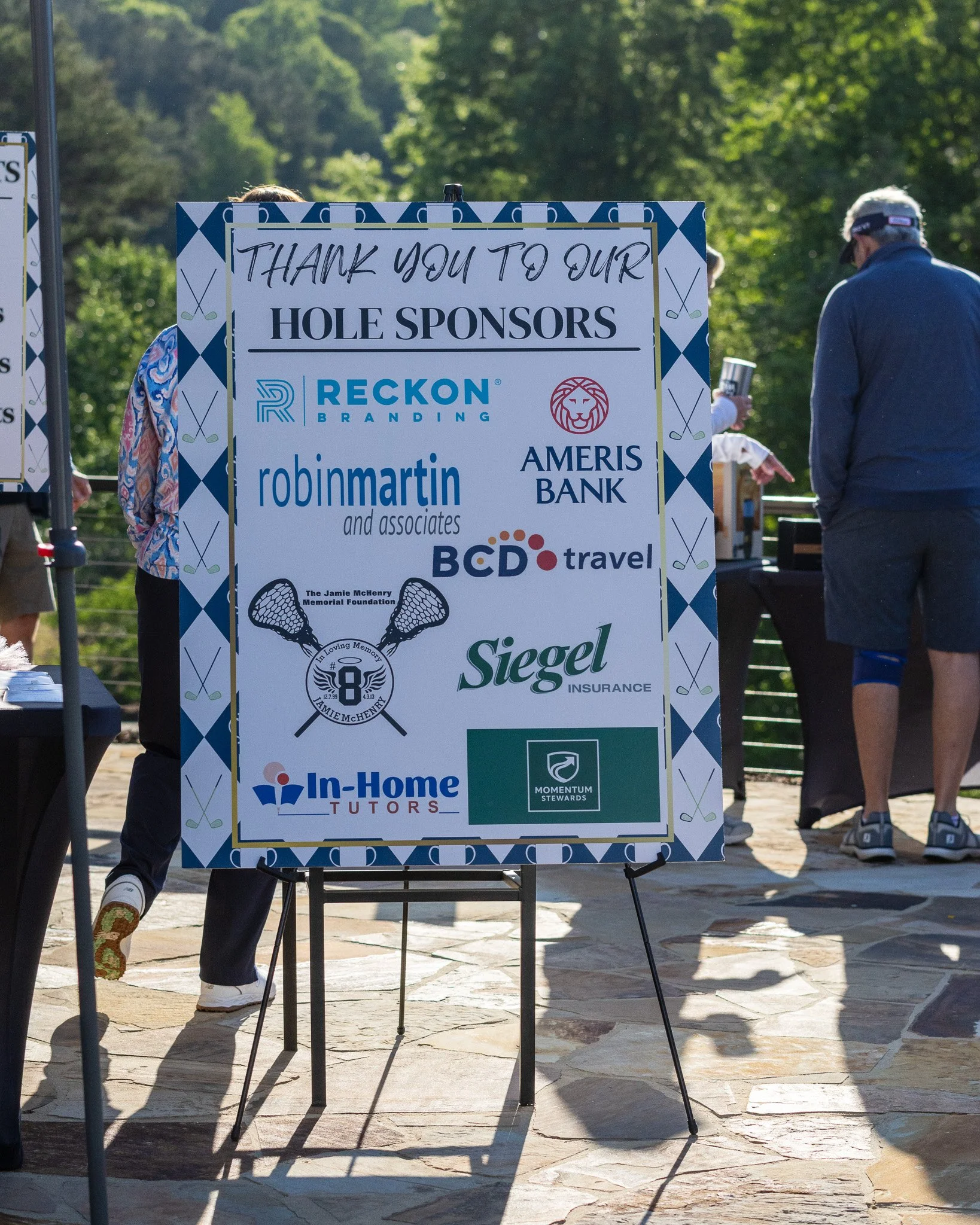 A large sign thanking hole sponsors for an event, featuring various sponsor logos, including Reckon Branding, Ameris Bank, Robin Martin and Associates, BCD Travel, Siegel Insurance, and In-Home Tutors, with people walking in the background outdoors o