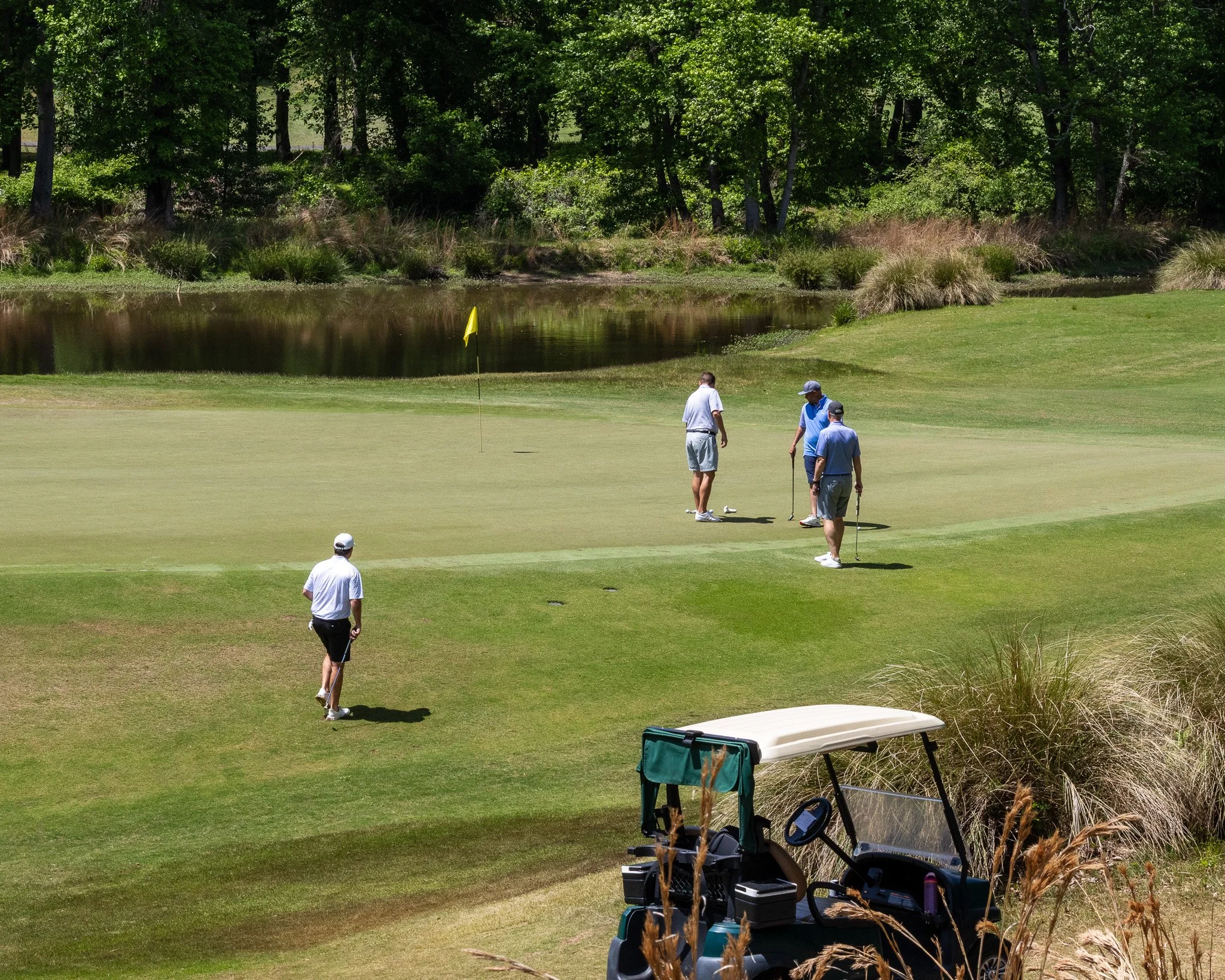 A group of men playing golf on a green course surrounded by trees and a pond, with golf carts and equipment nearby.