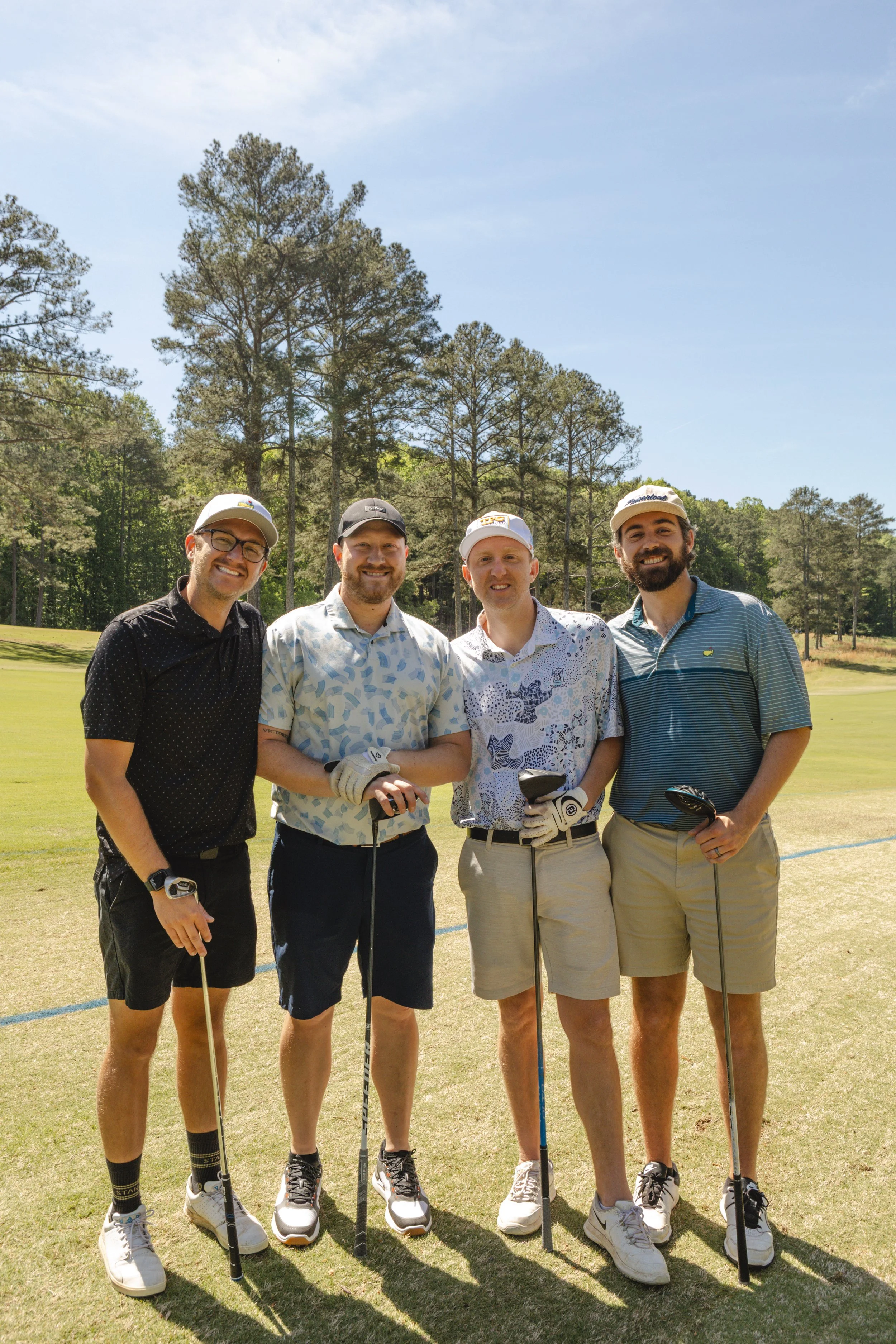Four men standing together on a golf course, holding golf clubs, smiling, with trees and a blue sky behind them.