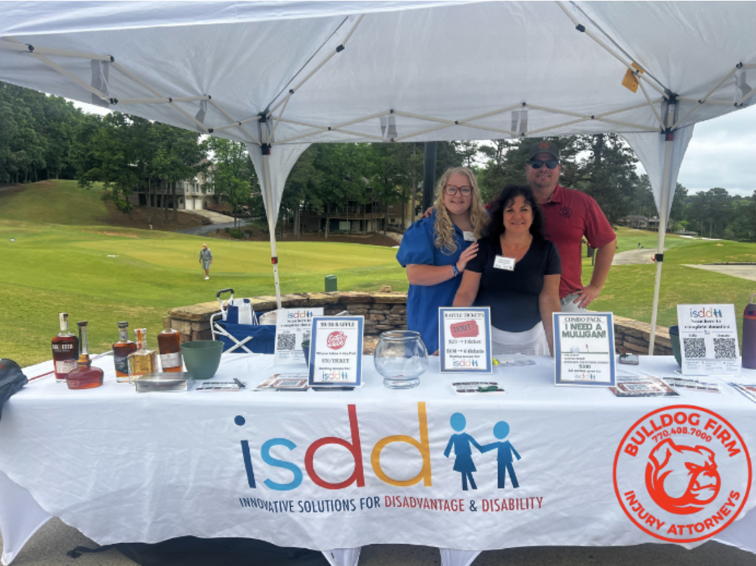 Three people standing behind a table with informational materials and drinks at an outdoor event, under a white canopy, with a golf course in the background.