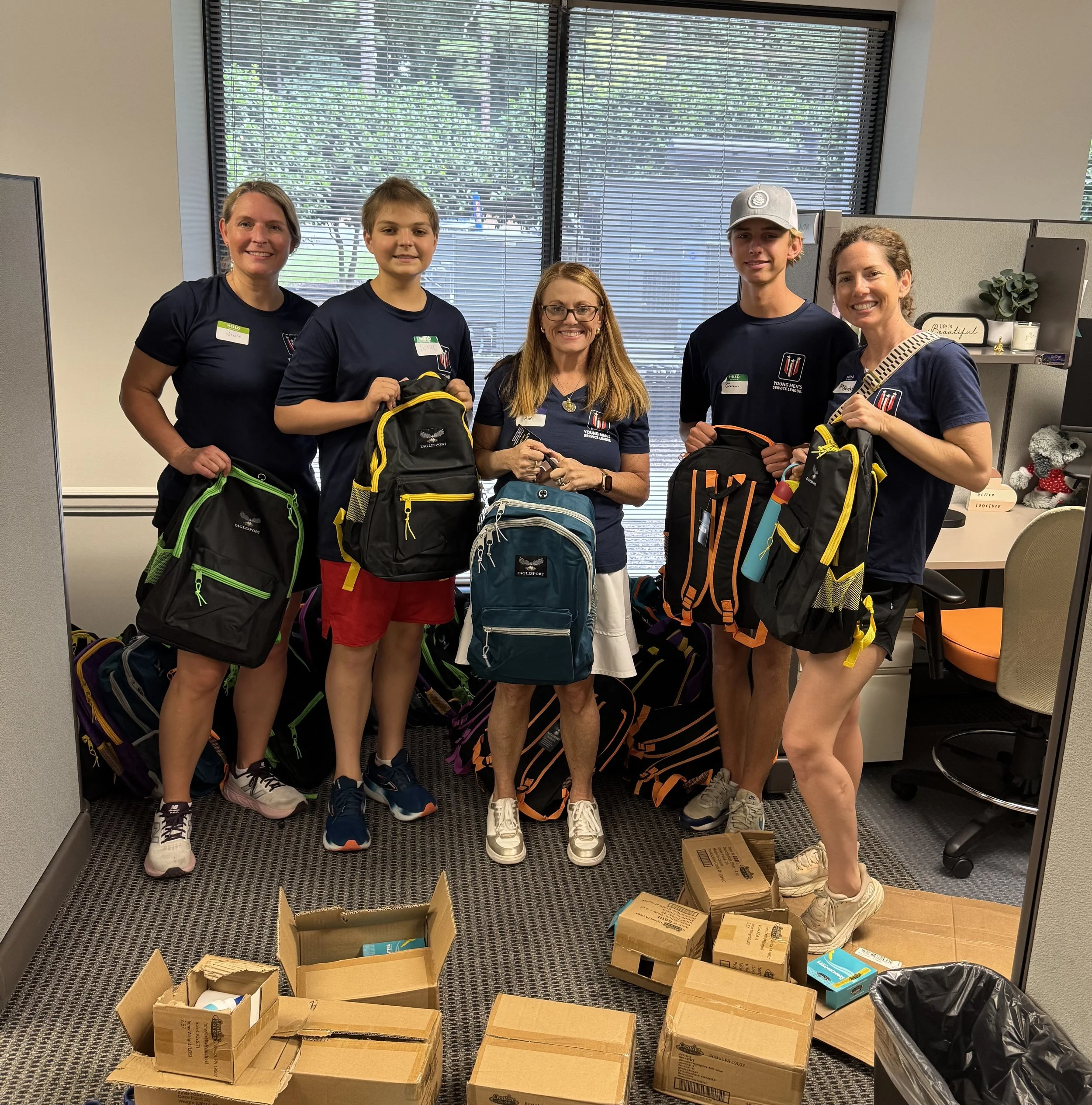 Five people standing inside an office, holding backpacks and surrounded by boxes. The group includes three women and two young men, all smiling and wearing dark t-shirts with event logos. The office has large windows with blinds, and there are some d