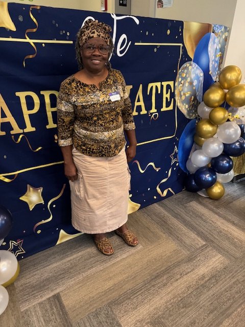 A smiling woman in a leopard print top and light skirt standing in front of a birthday backdrop with balloons.
