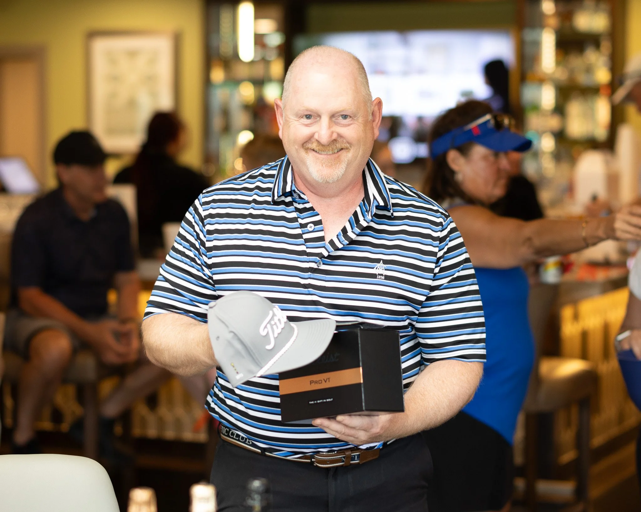 A smiling man with a goatee and blue eyes wearing a striped blue, white, and black shirt standing in a busy indoor space, holding a boxed golf glove. There are blurred people in the background, including women working or shopping, and shelves with va