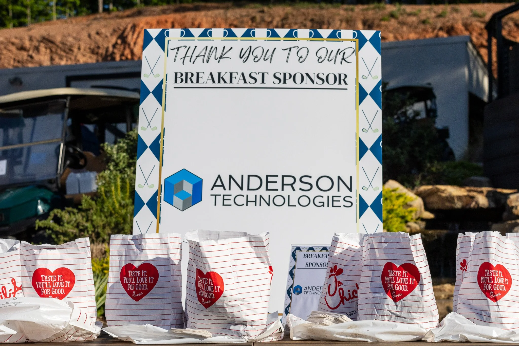A large outdoor sign thanking a breakfast sponsor named Anderson Technologies, with a row of Chick-fil-A paper bags with red hearts and messages of appreciation in the foreground.
