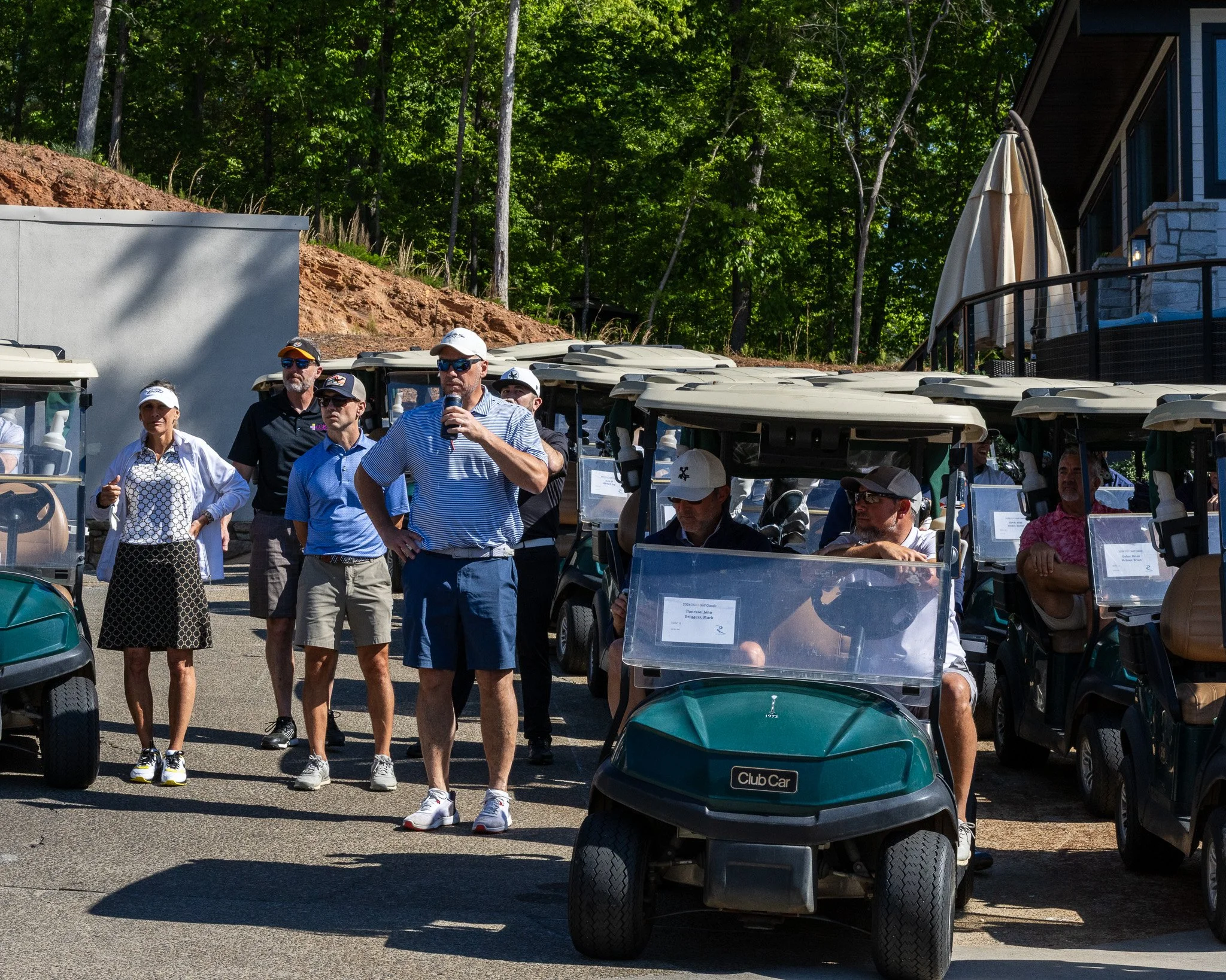 Group of people at a golf course, some standing and some seated in golf carts, with trees and a building in the background.