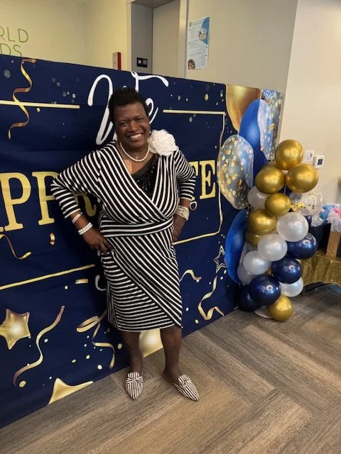 A woman in a black and white striped dress standing in front of a navy blue and gold congratulatory backdrop, with a balloon arrangement of blue, white, gold, and navy balloons to her right.