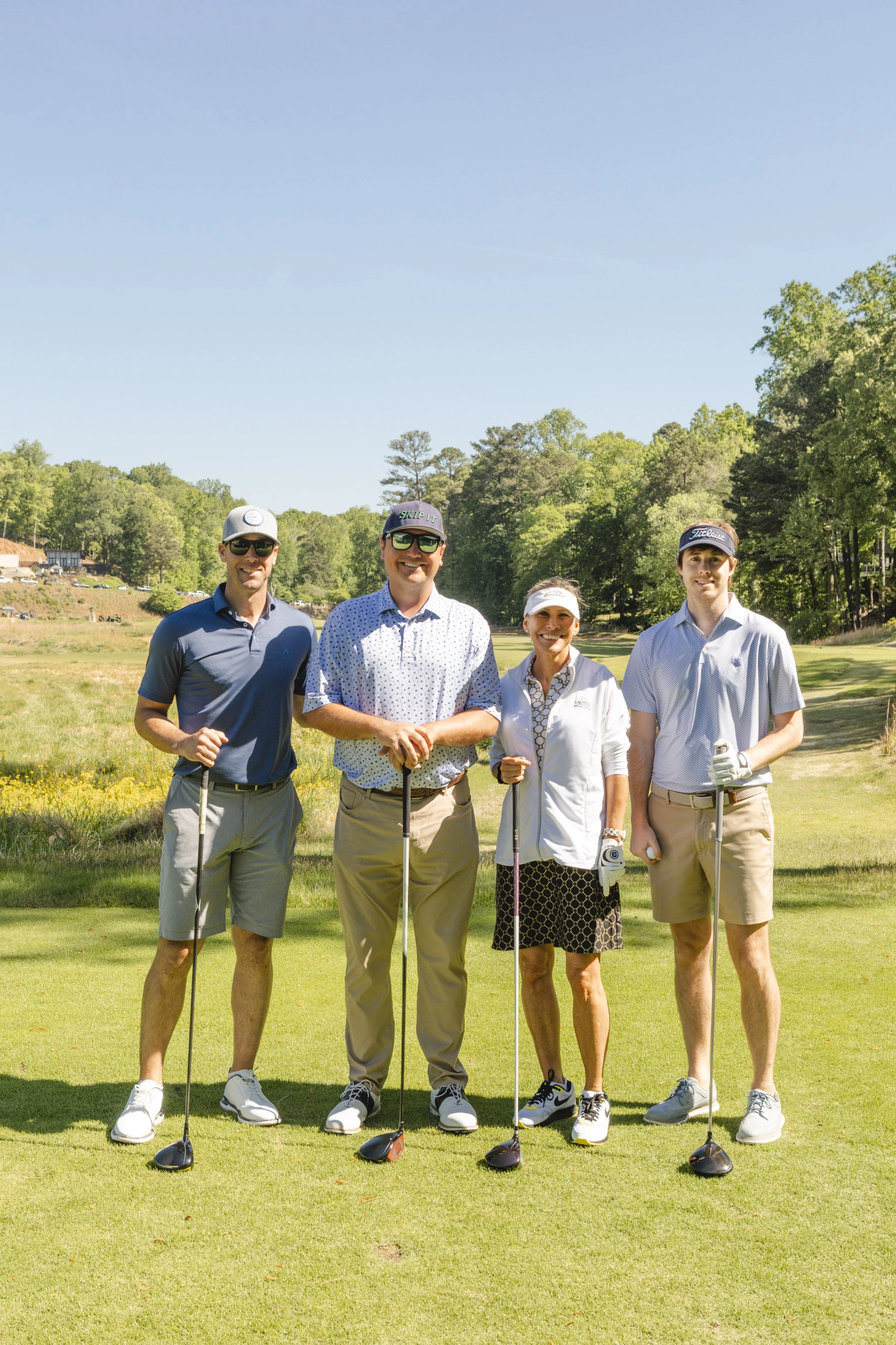 Five people standing on a golf course, smiling, holding golf clubs, with green trees and a blue sky in the background.