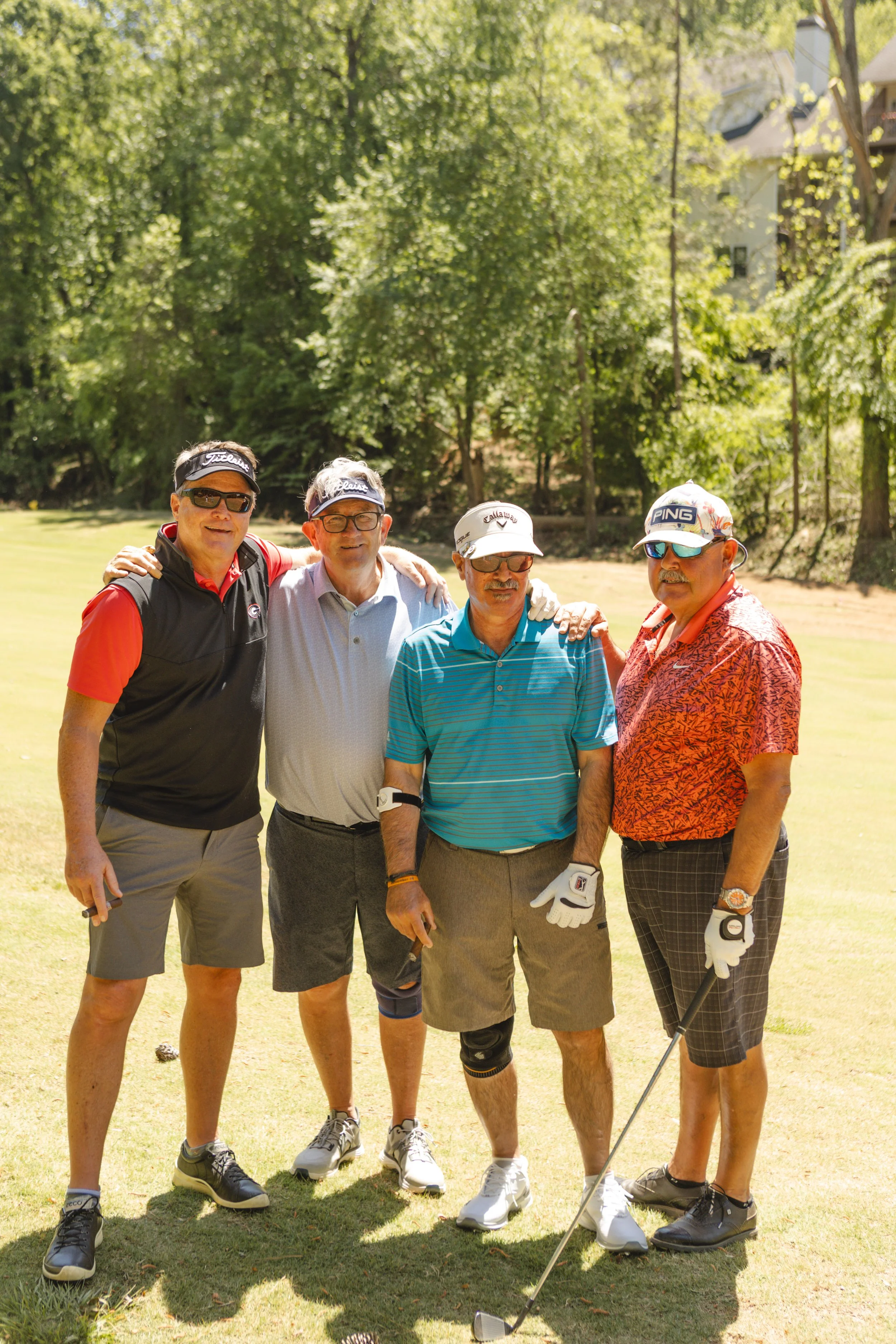 Four older men standing on a golf course, dressed in golf attire, holding golf clubs, smiling, with trees and a white house in the background.