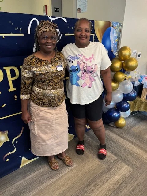 Two women standing indoors near a balloon display with gold, navy blue, and white balloons, and a decorated backdrop.