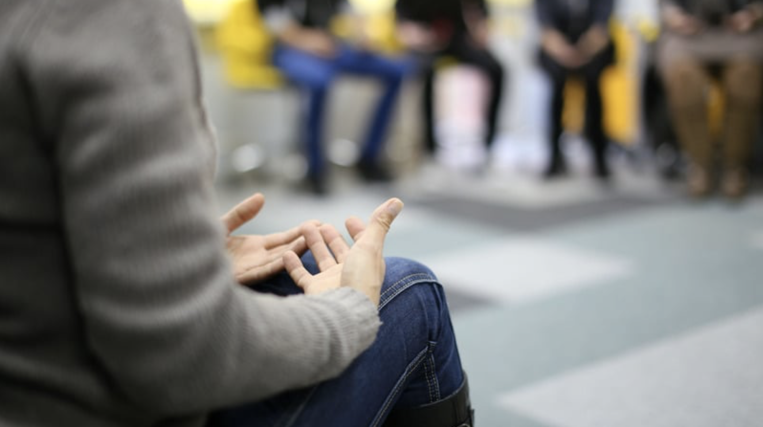 Person wearing a gray sweater and blue jeans sitting with hands gesturing, in a busy public space with blurred people sitting on yellow seats in the background.
