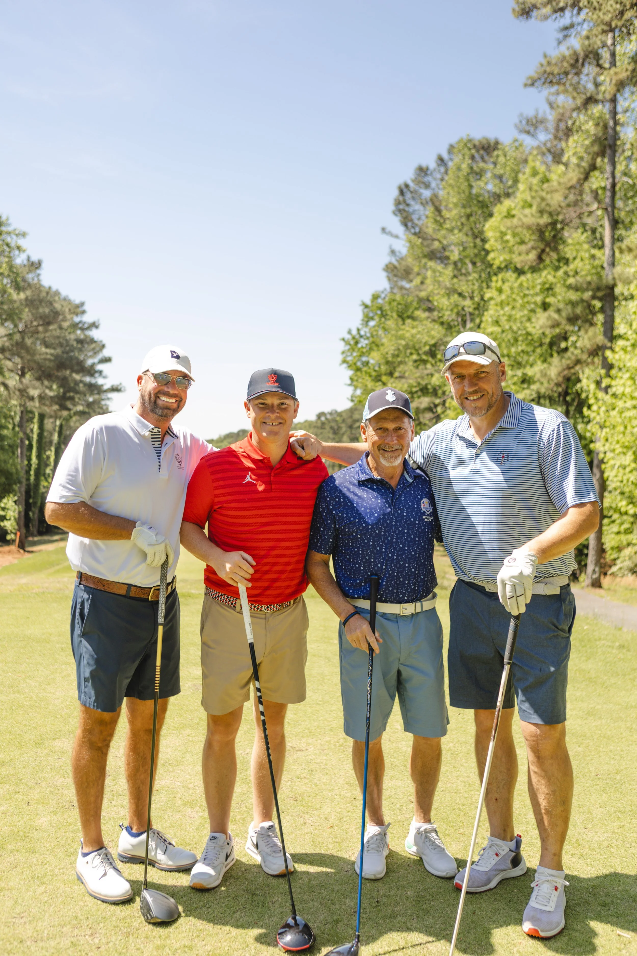 Five men standing on a golf course, smiling, holding golf clubs, wearing golf attire, with trees and blue sky in the background.