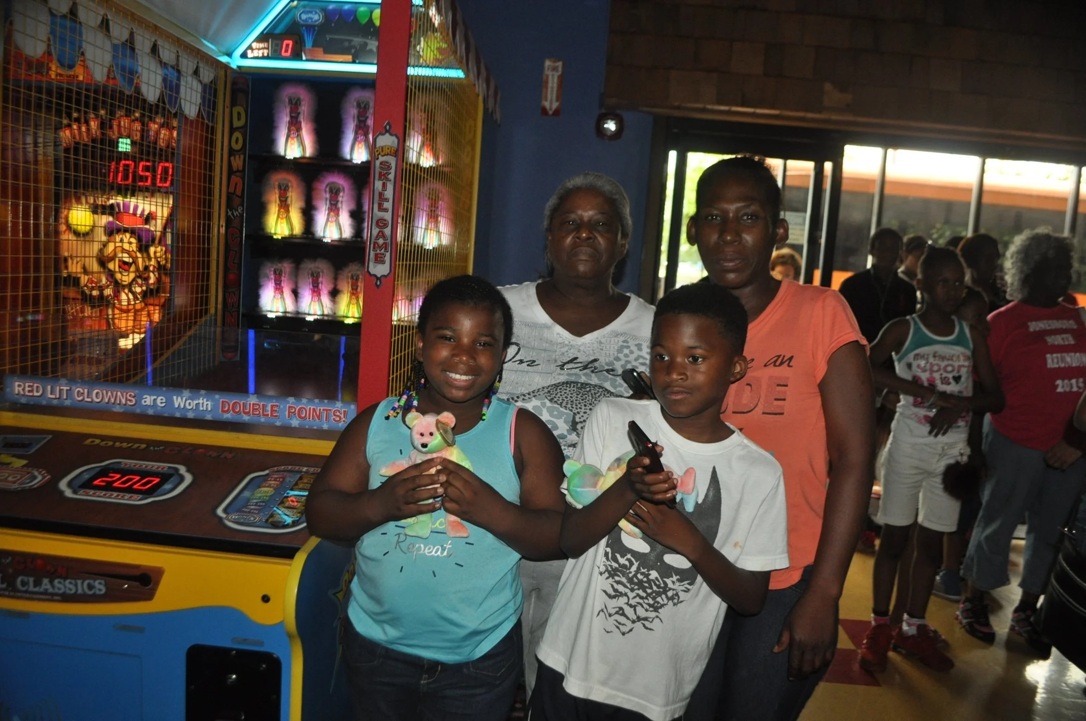 Three people, two women and one girl, standing in front of an arcade game at a lively amusement center. The girl is smiling and holding stuffed animals, while the women stand behind her. Other people are visible in the background.