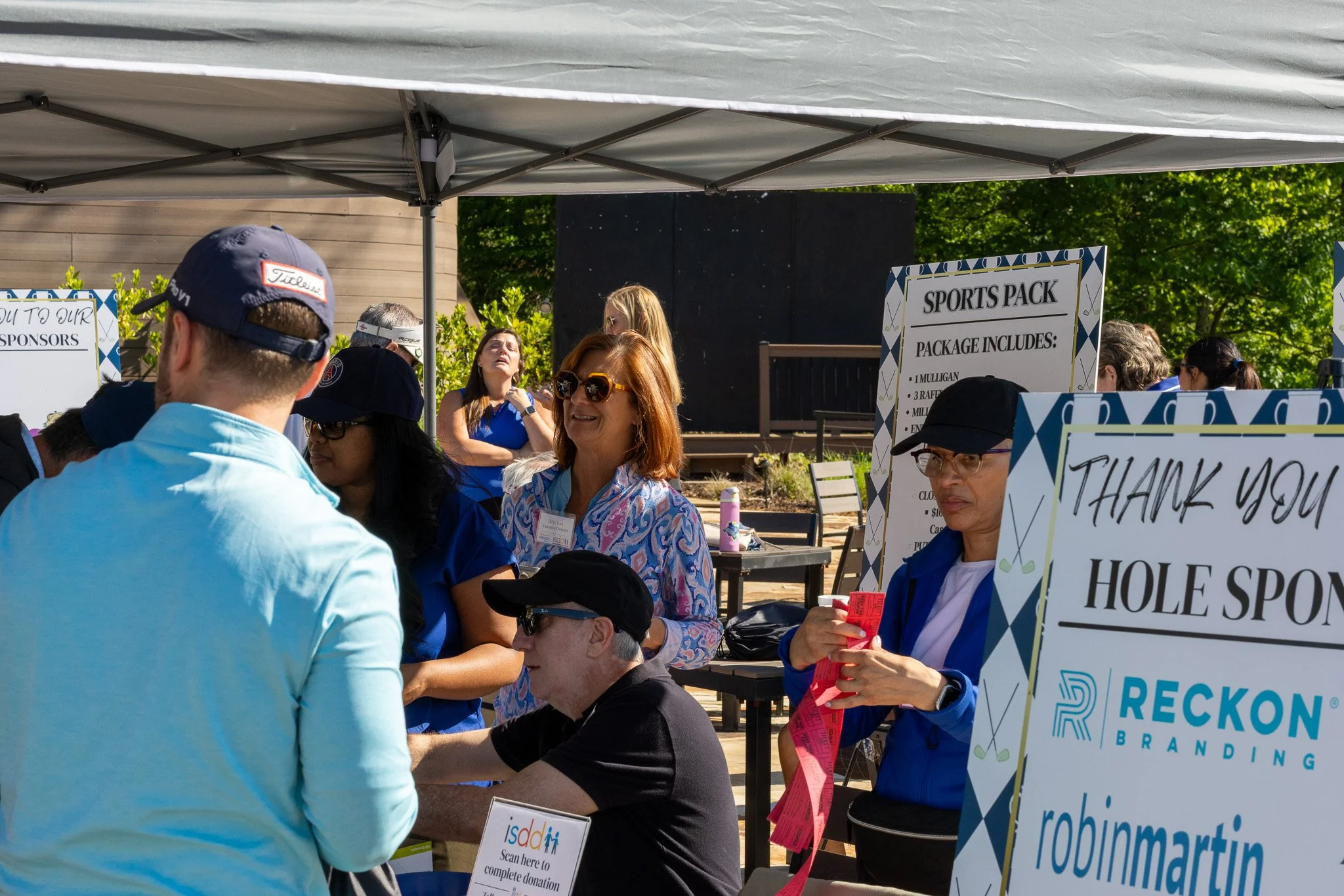 People gathered at an outdoor event, some wearing hats and sunglasses, with banners and signs advertising sponsorships and donations, under a canopy with trees in the background.