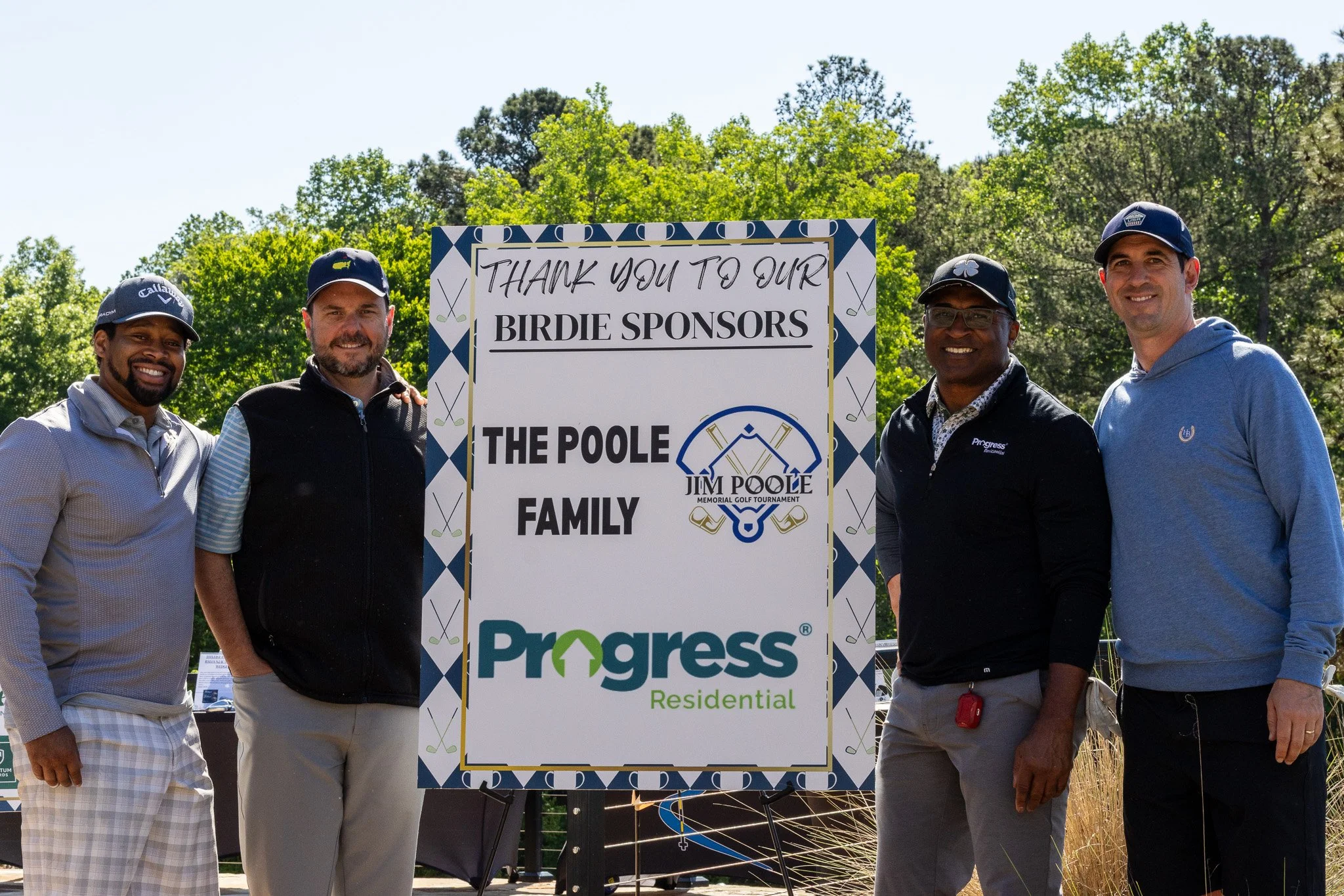 Group of five men standing outdoors behind a sign that reads 'Thank you to our birdie sponsors, The Poole Family' at a golf event, with trees and blue sky in the background.