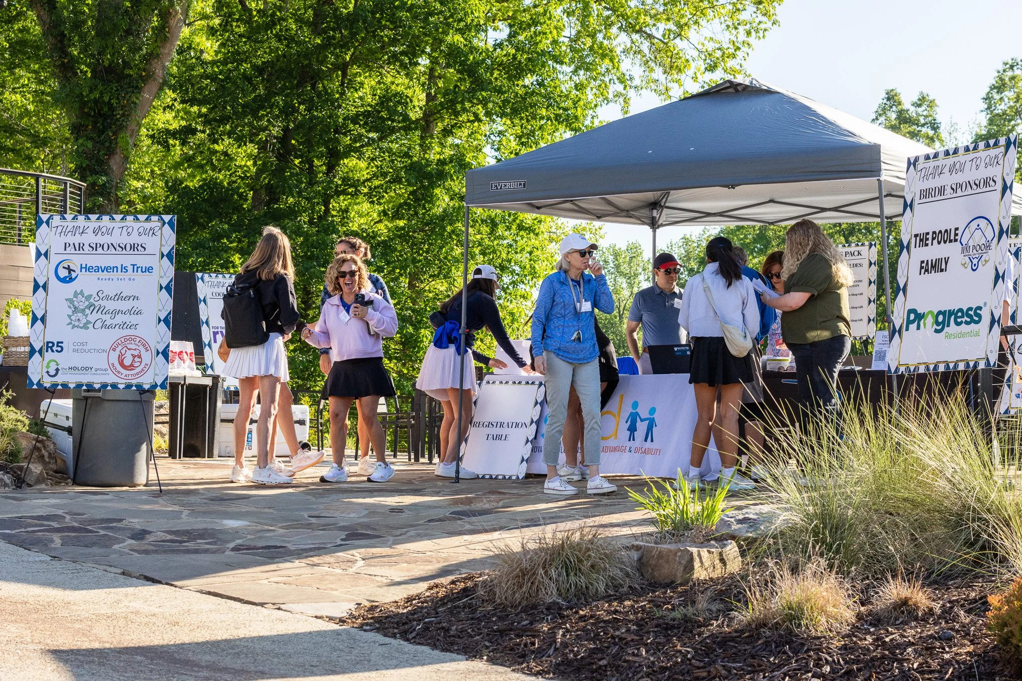 People gather at an outdoor event or festival, standing under a canopy with banners and signs thanking sponsors, surrounded by trees and greenery, sunny weather.